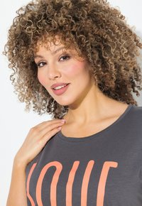 Camiseta gris con grandes letras naranjas que dicen "AMOR". El tejido parece suave con una textura lisa y un escote redondeado.