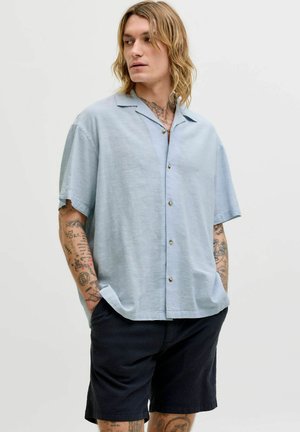 Mann mit Tätowierungen, der ein hellblaues kurzärmeliges Hemd mit Knöpfen und schwarze Shorts trägt und mit den Händen in den Taschen steht.