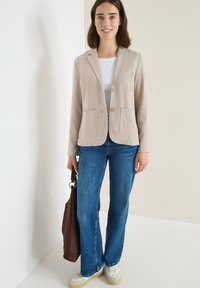 Beige gestructureerde blazer met twee voorzakken, gedragen over een witte top, gecombineerd met blauwe flared jeans en crème sneakers, met een bruine tas in de hand.