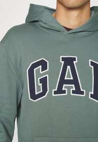 Felpa verde con cappuccio con un grande logo "GAP" blu navy contornato di bianco. Il tessuto sembra morbido con una vestibilità standard e tasca frontale.