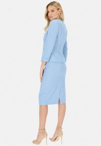 Niebieski garnitur z tweedu, z dopasowaną marynarką i spódnicą do kolan, z rozcięciem z tyłu i rękawami do trzyczwartych. Wysokie obcasy dopełniają stylizację.