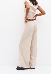 Kobieta poprawiająca talię beżowych, prążkowanych spodni szerokich nogawkach, w połączeniu z bezrękawnikami oraz ciemnobrązowymi butami na platformie, na tle białego tła.
