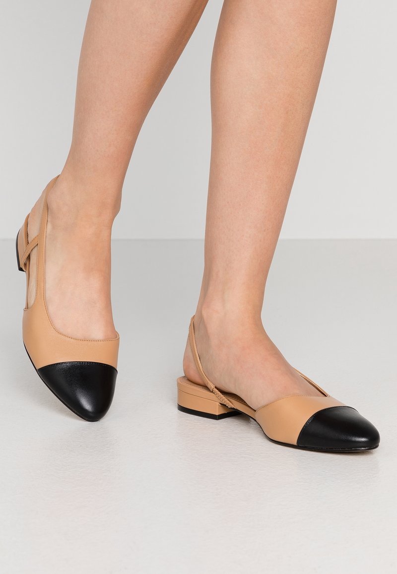 Beige og sorte slingback lavhælede sko båret på barfod mod en enkel lys baggrund.