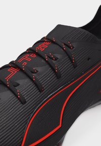 Μαύρο αθλητικό παπούτσι με κόκκινες λεπτομέρειες, εκλεπτυσμένα κορδόνια, υφή στην πάνω επιφάνεια και εμφανές λογότυπο στη γλώσσα και στην πλευρική επιφάνεια.