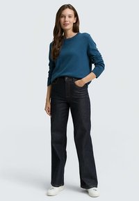 Žaliai mėlynas megztinis su ilgomis rankovėmis, derinamas su tamsiai mėlynais tiesia kojų linija džinsais ir baltais sportbačiais. Paprastas dizainas, laisvas siluetas ir vientisos spalvos.