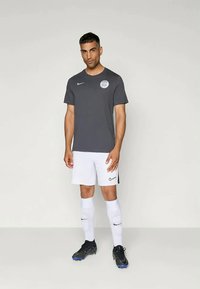 T-shirt Nike gris à manches courtes avec un logo circulaire, associé à un short blanc et des chaussettes de football. Chaussons noires avec des accents bleus.