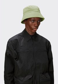 Chapeau seau vert clair avec une texture côtelée. Porté par une personne dans une veste noire zippée avec une finition froissée et deux poches zippées.