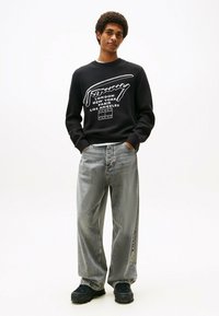 Schwarzer Pullover mit weißem Drucktext, kombiniert mit lockeren grauen Jeans und schwarzen klobigen Schuhen, vor einem schlichten weißen Hintergrund.