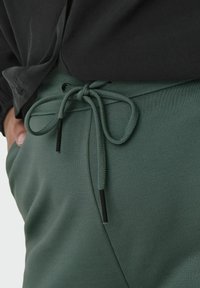 Grønne joggebukser med snøring i livet, med en glatt tekstur og svart metall på snøringen. En del av en mørk topp er også synlig.