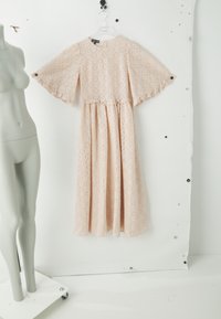 Vestido de encaje rosa pálido con mangas cortas de vuelo, patrón geométrico, cuello redondo y falda fruncida colgado en una percha transparente sobre un fondo blanco.