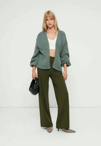 Pilkai žalsvas megztinis su nuleistais pečiais, dėvimas virš balto trumpinto viršaus ir derinamas su alyvuogių spalvos plačiomis kelnėmis bei smėlio spalvos aukštakulniais.