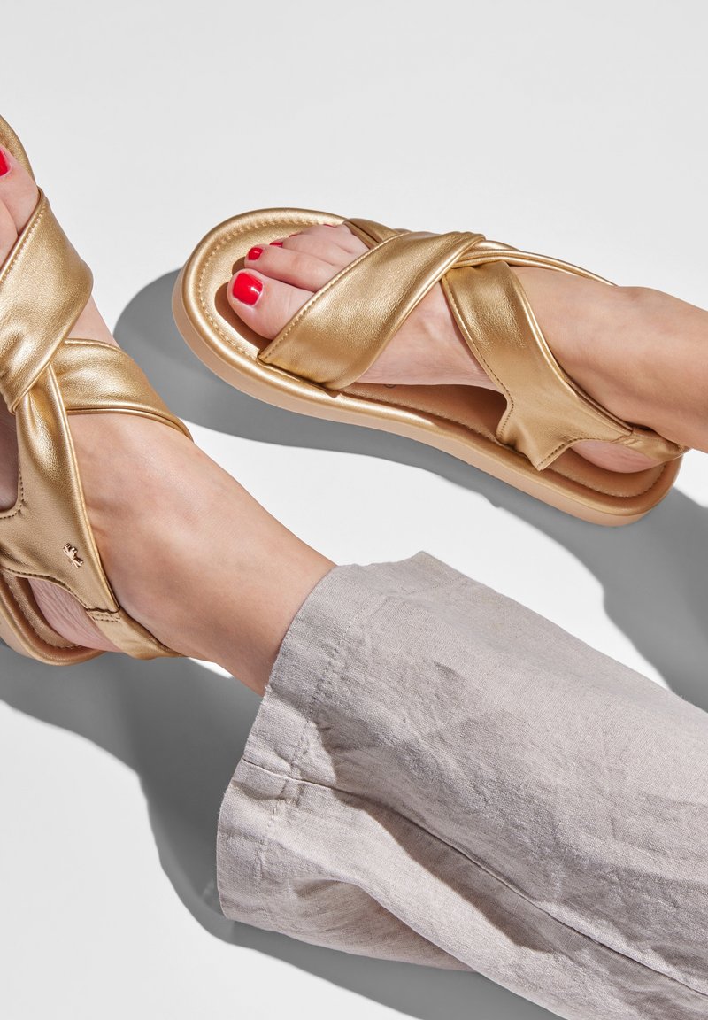 Gold cross-strap sandals feature a shiny metallic finish, soft padded sole, and a lightweight construction. Paired with light linen pants.