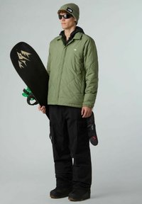 Uomo con berretto verde, occhiali da sole e giacca trapuntata verde che tiene una tavola da neve nera con un design montano, vestito con pantaloni da neve neri e stivali.