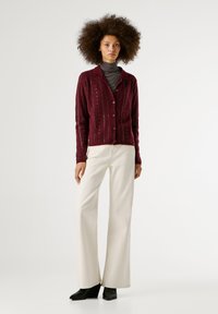 Μπούρντι υφασμένο γιλέκο με κουμπιά μπροστά, υφές και σχέδιο γιακά, συνδυασμένο με κρεμ παντελόνια καμπάνα και μαύρα παπούτσια.