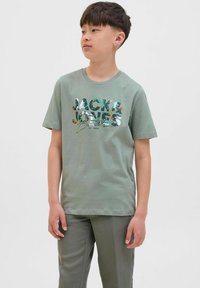 Svijetlozeleni pamuk majica s kratkim rukavima i grafičkim tiskom "JACK & JONES" u raznolikim bojama. Ima okrugli izrez i ležeran kroj.