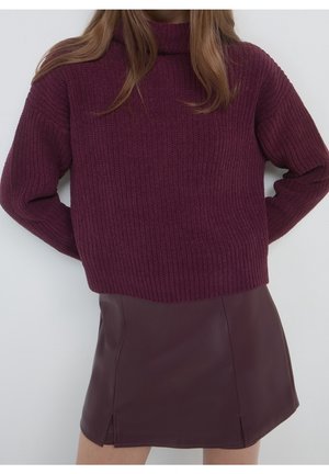 Femme portant un pull en grosse maille bordeaux et une mini-jupe en cuir assortie, debout les mains derrière le dos.