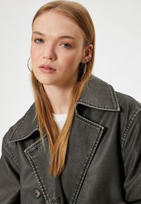 Veste en faux cuir vert foncé avec de grands revers et une fermeture à double bouton. Le mannequin a les cheveux longs et raides ainsi que de petites boucles d'oreilles en anneau.