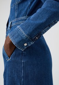 Denimklänning med en figursydd design i mellanblått, långa ärmar med knappslut och sidofickor. Synlig sömnad och tygtextur.