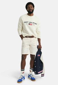 Crèmekleurig sweatshirt met de tekst "GANT USA SPORT", beige shorts, blauwe sneakers en een marineblauwe jack over de arm. Accessoires zijn witte sokken.