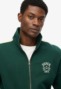 Grønne zippered sweatshirt med cremefarvede detaljer, prydet med et "Super Dry" logo i hvidt og en tekstureret stof. Modellen har en hvid T-shirt på under.