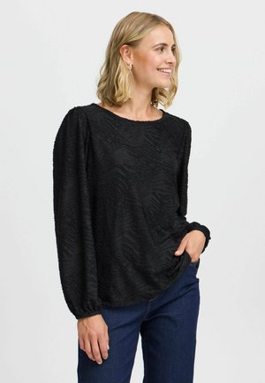 Sort tekstureret bluse med lange ærmer og rund halsudskæring. Har et bølget mønster og elastiske manchetter. Kombineret med mørkeblå jeans.