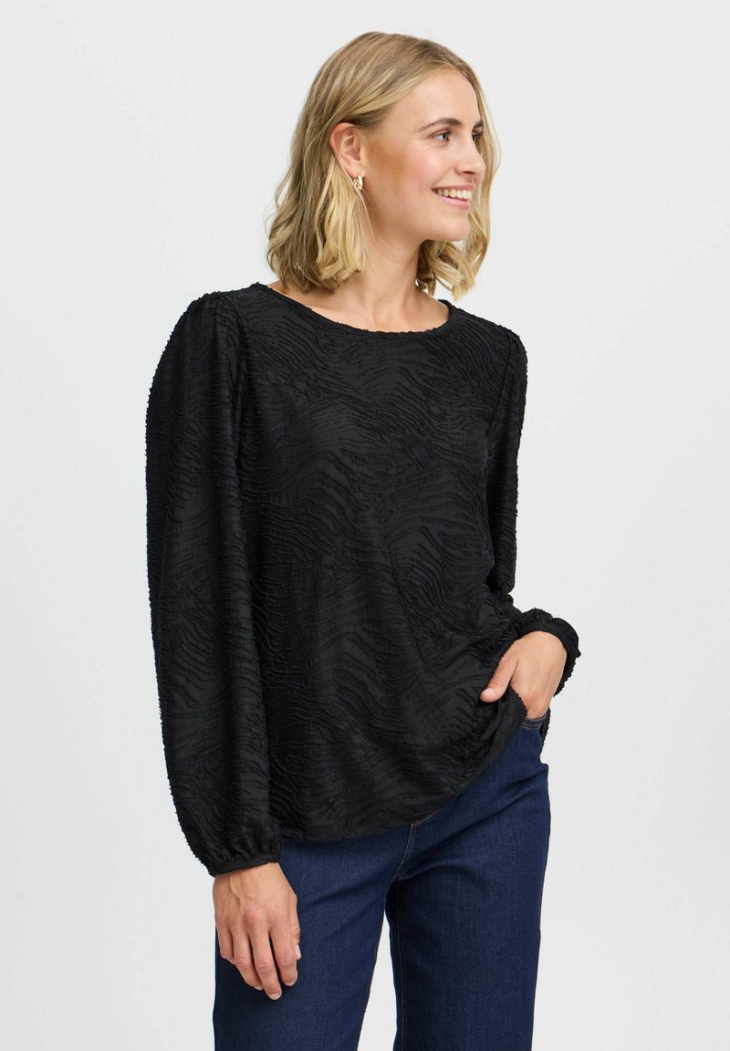 Schwarze strukturierte Bluse mit langen Ärmeln und Rundhalsausschnitt. Merkmale ein welliges Muster und elastische cuffs. Kombiniert mit dunkelblauen Jeans.
