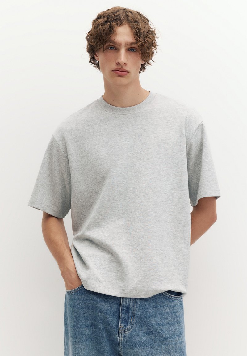 Jeune homme aux cheveux bouclés portant un t-shirt ample gris clair et un jean bleu, debout les mains dans les poches devant un fond uni.