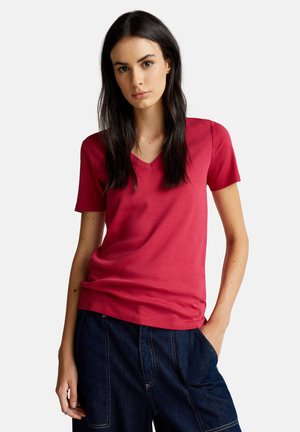 Vrouw met lang donker haar die een rode T-shirt met korte mouwen en een V-hals draagt, en donkere blauwe spijkerbroek, poseert met één hand in de broekzak.