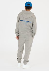 Grauem Sweatshirt und Jogginghosen aus Baumwollmaterial. Das Sweatshirt hat auf der Rückseite den blauen Text „NEW BAD HABITS“; die Jogginghosen haben einen Bündchenabschluss. Weiße Turnschuhe.