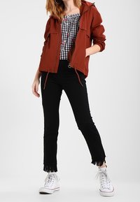 Chaqueta con capucha de color óxido con bolsillos, top a cuadros en blanco y negro, vaqueros negros recortados con dobladillos deshilachados y zapatillas blancas.