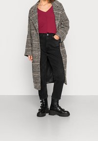 Černobíle kostkovaný dlouhý kabát s širokými rukávy přes vínový top, doplněný o černé džíny s vysokým pasem a robustní černé boty.
