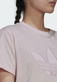 Světle růžové bavlněné tričko s kulatým výstřihem, s vyšitým logem na hrudi. Krátké rukávy a jemná textura.