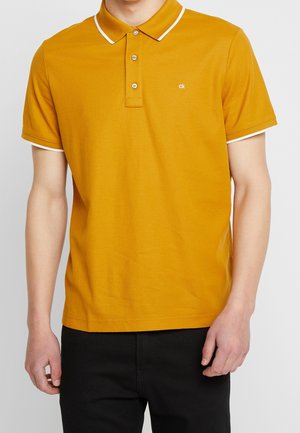 Mustard polo shirt with a white collar and sleeve trims, three-button placket, and small logo on the chest. Cotton material.