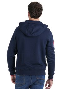 Sweatshirt à capuche bleu marine en mélange de coton. Présente des poignets et un ourlet côtelés, avec un patch logo sur la manche gauche. Texture lisse.