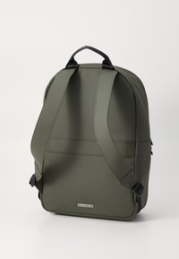 Mochila en color verde oliva con un acabado liso y mate, que incluye correas acolchadas, un asa en la parte superior y una placa de metal con el logo en el frente.