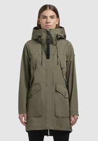Parka en vert olive avec capuche à cordon de serrage, zip à l'avant, poches à pression et une étiquette logo audacieuse au niveau du cou. Tissu lisse et léger.