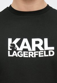 Melna kokvilnas T-krekls ar izteiksmīgu baltu tekstu "KARL LAGERFELD" un profila grafiku pa kreisi. Apaļš kakla izgriezums.