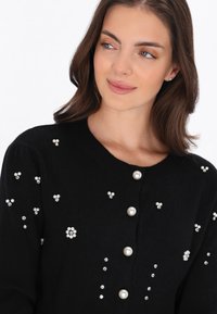 Svart ribbad cardigan med pärl- och kristalldetaljer, rundad halsringning och knäppning, med ett texturerat tyg och detaljerade accenter.