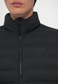 Primo piano di una persona che indossa un giaccone nero imbottito con colletto alto, cerniera visibile e logo EA7 Emporio Armani sul petto.