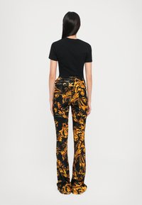 Camiseta negra combinada con pantalones negros acampanados que presentan un atrevido patrón barroco en amarillo y dorado, textura suave y silueta ajustada.