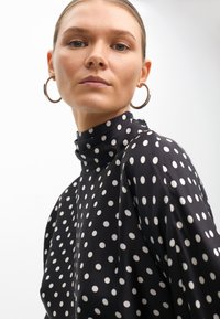 Schwarze Bluse mit Punkten, hohem Kragen und langen Ärmeln, versehen mit großen weißen Punkten und einer glatten, satinähnlichen Textur.