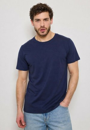 Homme aux cheveux courts et bouclés avec des poils du visage, portant un t-shirt bleu marine et un jean bleu clair, debout devant un fond blanc uni.