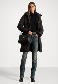 Geacă puffer neagră cu glugă, fermoar frontal și accent de logo. Purtată peste un pulover din lână închisă la culoare și blugi, asortată cu o poșetă neagră.