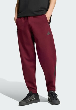 Person trägt burgunderrote Adidas Trainingshose mit Reißverschlusstaschen und schwarze Adidas Sneakers mit weißen Streifen, steht mit den Händen in den Taschen.