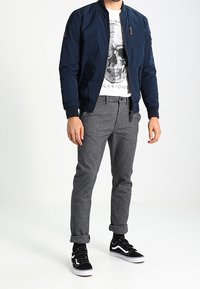Marine bomberjack, graphic T-shirt met schedelontwerp, grijze pantalon en zwarte sneakers met witte accenten en textielstof.