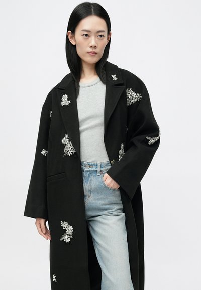 Črn plašč z vezenimi cvetličnimi vzorci v srebrni barvi, z zareznim ovratnikom, dolgimi rokavi in sproščenim krojem, v kombinaciji s sivo majico in svetlo modrimi kavbojkami.
