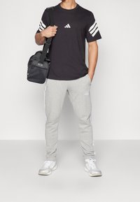 Černé tričko Adidas s bílým třípruhovým designem na rukávech, světle šedé tepláky s bílými bočními pruhy a šedé sportovní boty.