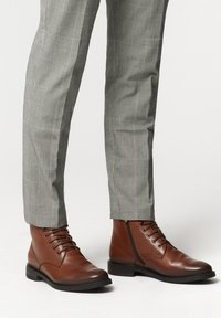 Brązowe skórzane botki za kostkę o gładkim wykończeniu, okrągłym nosku i sznurowanym designie. W komplecie z szarymi spodniami w kratę, mającymi teksturowany materiał.