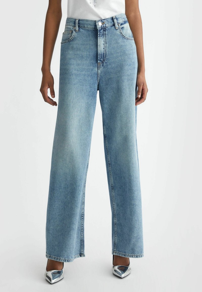 Personne portant un jean large bleu clair et des chaussures à talons aiguilles argentées, debout devant un fond uni.