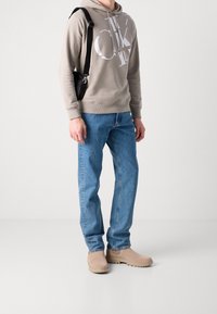 Sudadera beige con gran logo blanco, jeans rectos azules y botas beige sin cordones con suelas texturizadas. Incluye bolso cruzado negro.